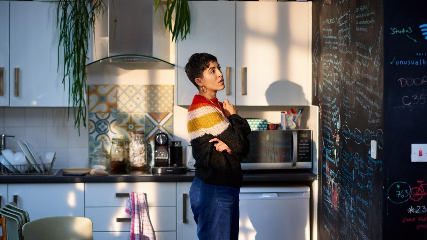 Woman standing in kitchen looks confused at blackboard covered in notes.