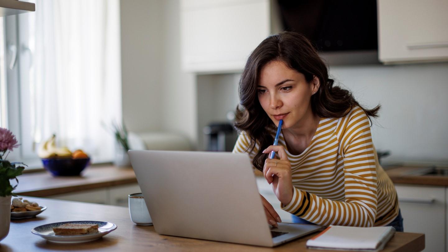 woman looking at a laptop in her kitchen, holding a pen to her mouth