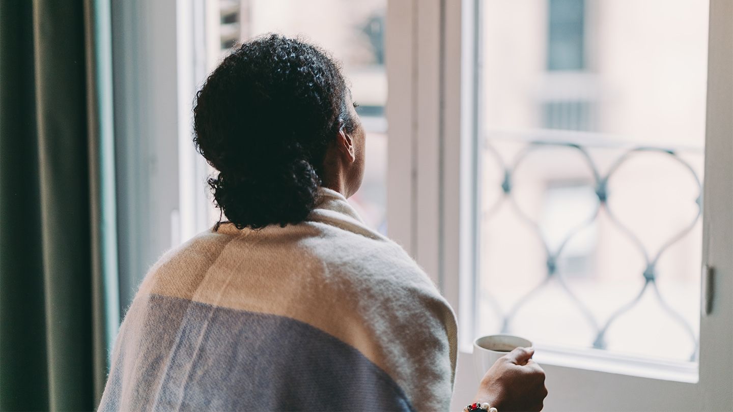 A woman looks out her window, holding a mug. 