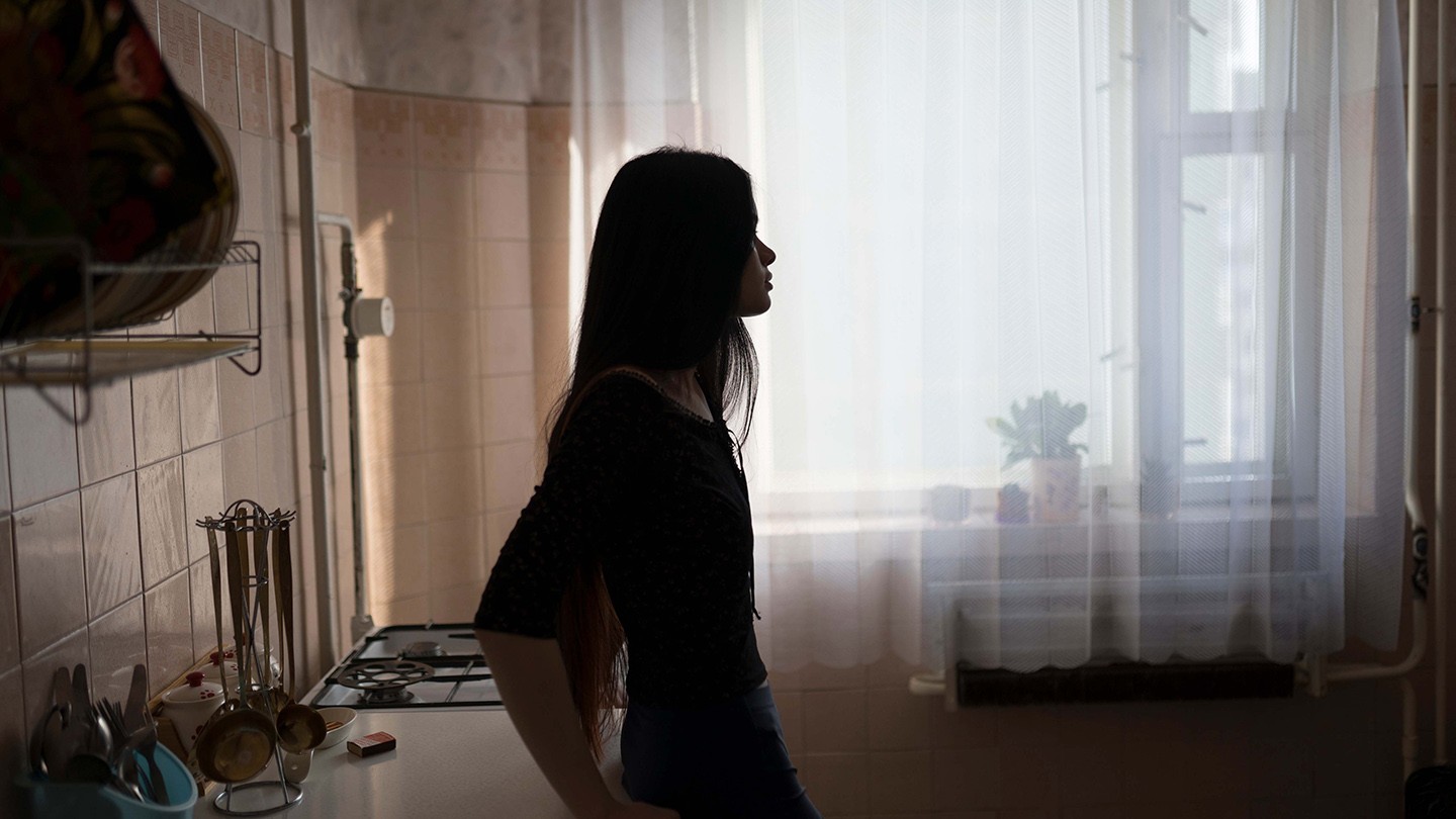 A woman leans on a kitchen counter and is silhouetted against a window. 
