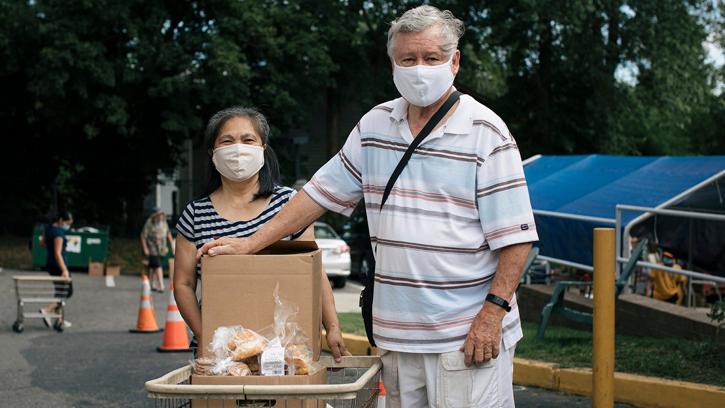 Two people wearing masks stand with a cart of food.