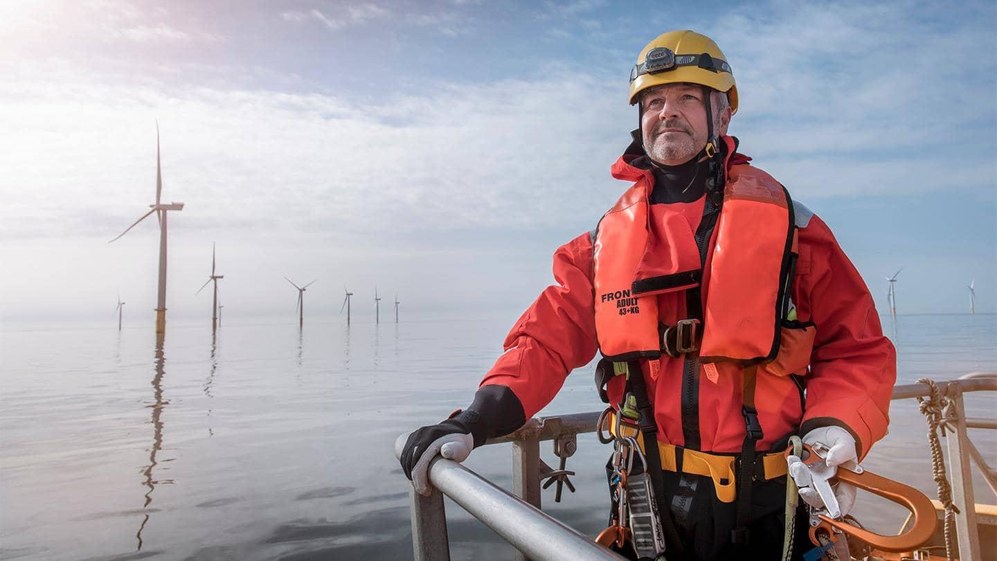 A man wearing a life jacket and helmet, standing by water with wind turbines.