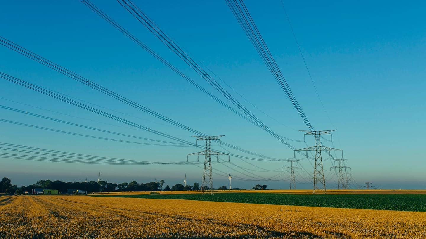 Two rows of electricity pylons in a yellow and green farmers field, with a blue sky. Wind turbines and trees are visible in the distance.